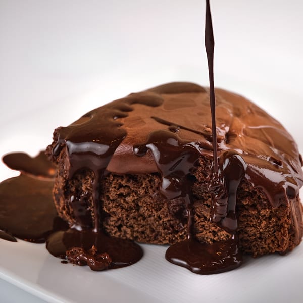 Rich chocolate sauce pours over a vegan chocolate cake with chocolate frosting on a white plate.