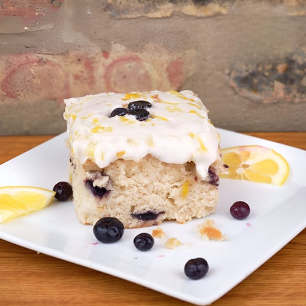 A slice of vegan lemon blueberry cake with white frosting and lemon zest on a white plate.