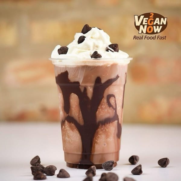 Vegan Now 2 Go chocolate milkshake with whipped cream and chocolate chips, against a brick wall.